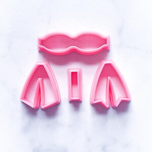 May include: A set of four pink plastic cookie cutters shaped like a bow tie, two bow ties, and a rectangle.