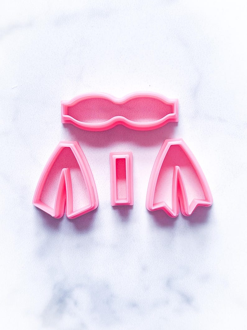 May include: A set of four pink plastic cookie cutters shaped like a bow tie, two bow ties, and a rectangle.