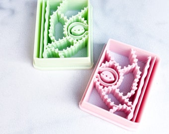 Puede incluir: Dos cortadores de galletas rectangulares, uno verde claro y otro rosa, se muestran sobre una superficie blanca. Cada cortador presenta un diseño intrincado con un elemento circular central y un patrón circundante. Los cortadores probablemente se utilizan para hornear.