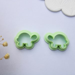 May include: Two light green turtle-shaped cookie cutters. The cutters are made of plastic and have a layered design. The image also includes a white decorative element and dried flowers, suggesting a crafting or baking theme.