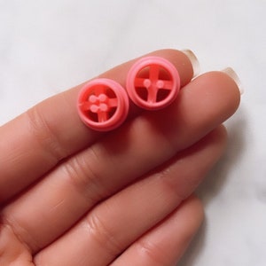 May include: Two pink plastic dollhouse furniture pieces. Each piece is a round, flat, circular shape with a raised design in the center. The pieces are similar in size and shape.