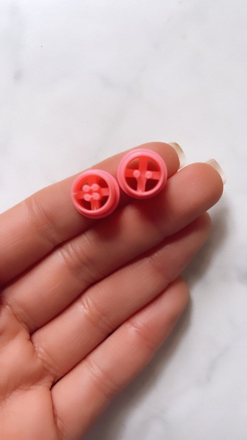 May include: Two pink plastic dollhouse furniture pieces. Each piece is a round, flat, circular shape with a raised design in the center. The pieces are similar in size and shape.
