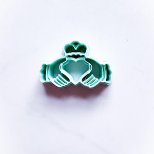 May include: A teal Claddagh cookie cutter. The design features a heart held by two hands, topped with a crown. The cutter is made of plastic and is designed for baking. The Claddagh is a traditional Irish symbol of love, loyalty, and friendship.
