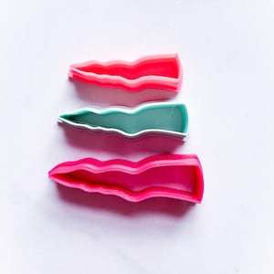 May include: Three colourful hair clips in shades of red, teal, and pink. Each clip has a wavy, textured design. The clips are arranged vertically against a white background, showcasing their vibrant colours and unique shape.