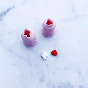 Puede incluir: Dos boquillas rosas con aberturas en forma de estrella y dos pequeñas decoraciones de fondant, un árbol blanco y un corazón rojo, sobre una superficie blanca. Herramientas para decorar pasteles.