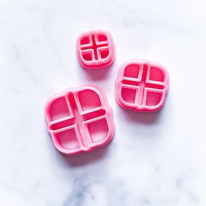 May include: Three pink plastic cookie cutters shaped like a square with a cross inside. The cutters are different sizes.