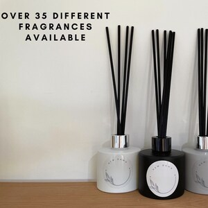 May include: Three diffuser bottles with black reeds. The bottles are white, black, and grey. Each bottle has a simple design with a circle and a leaf. The text "NEW DAWN" is on the black bottle. The text "OVER 35 DIFFERENT FRAGRANCES AVAILABLE" is at the top of the image.
