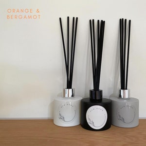 May include: Three glass diffuser bottles with black reeds. The bottles are labelled "New Dawn" and have a white circle with a leaf design. The scent is "Orange & Bergamot".