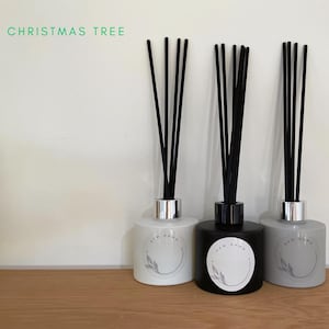 May include: Three reed diffusers with black reeds. The diffusers are in white, black, and grey glass bottles with silver caps. Each bottle has a white circle label with the text "NEW DAWN" and a small leaf design.