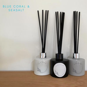 May include: Three glass diffuser bottles with black reeds. The bottles are labelled "New Dawn" and have a white, black, and grey colour scheme. The text "BLUE CORAL & SEASALT" is in the top left corner of the image.
