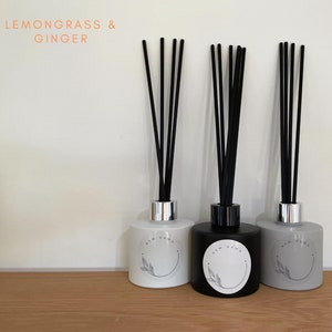 May include: Three reed diffusers with black reeds. The diffusers are in white, black, and grey glass bottles. Each bottle has a silver or black collar and a label with the text "NEW DAWN" and a leaf design. The text "LEMONGRASS & GINGER" is at the top.