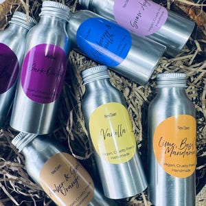 May include: A collection of eight small silver aluminium bottles with labels featuring different scents. The labels are round and feature a variety of colours, including purple, blue, pink, yellow, and orange. The labels also include the text "New Dawn" and the scent name. The bottles are arranged in a basket filled with shredded paper.