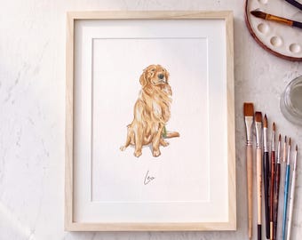 Puede incluir: Una ilustración en acuarela enmarcada de un perro golden retriever sentado, con el nombre Leo escrito en letra cursiva debajo del perro. La obra está en un marco de madera clara sobre una superficie blanca con pinceles.