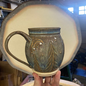 Puede incluir: Taza de cerámica hecha a mano con un asa grande. La taza presenta un esmalte azul oscuro y marrón con detalles dorados. El diseño incluye árboles estilizados y una luna creciente. La taza está sostenida en la mano de una persona.