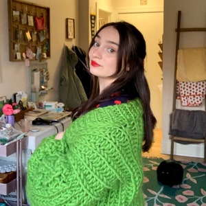 K&ouml;nnte beinhalten: Eine Frau tr&auml;gt einen leuchtend gr&uuml;nen Strickcardigan mit grober Strickstruktur. Der Cardigan hat lange &Auml;rmel und eine lockere Passform. Darunter tr&auml;gt sie ein Oberteil mit marineblauem und rotem Muster.