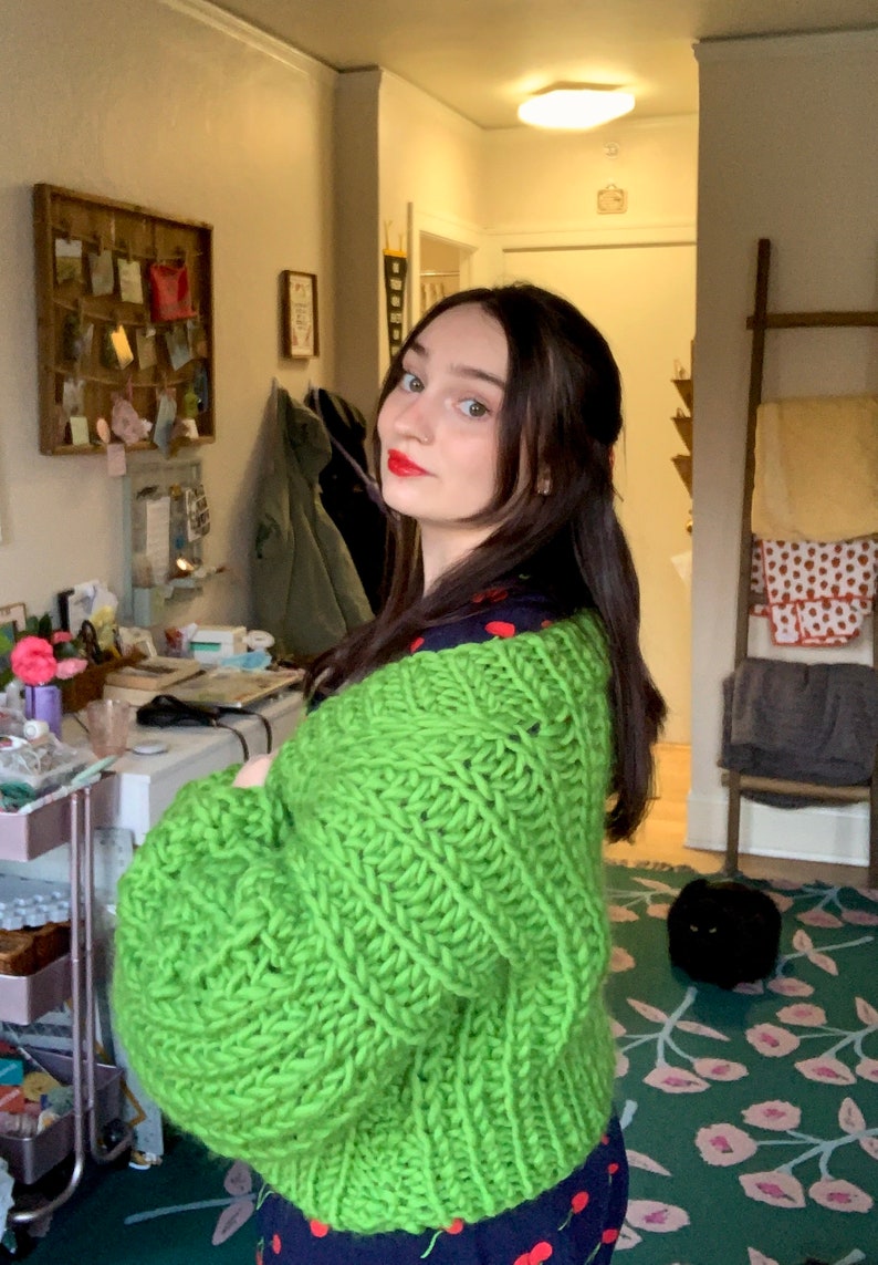 K&ouml;nnte beinhalten: Eine Frau tr&auml;gt einen leuchtend gr&uuml;nen Strickcardigan mit grober Strickstruktur. Der Cardigan hat lange &Auml;rmel und eine lockere Passform. Darunter tr&auml;gt sie ein Oberteil mit marineblauem und rotem Muster.
