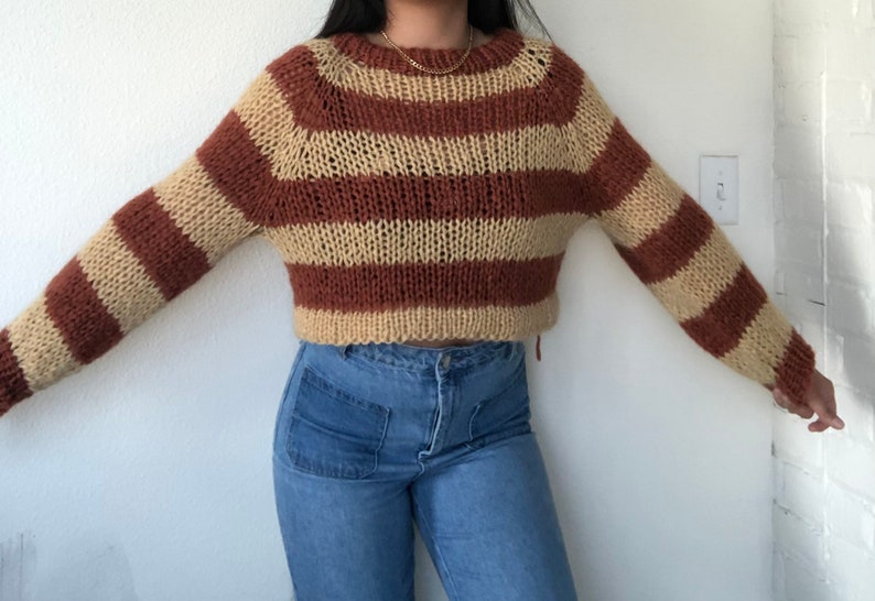 May include: A brown and beige striped knitted sweater with a cropped length. The sweater has a relaxed fit and a round neckline.