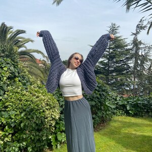 K&ouml;nnte beinhalten: Eine Frau tr&auml;gt einen grauen grob gestrickten Cardigan mit einem wei&szlig;en Crop-Top und einem langen grauen Rock. Sie steht in einem Garten mit Palmen im Hintergrund.