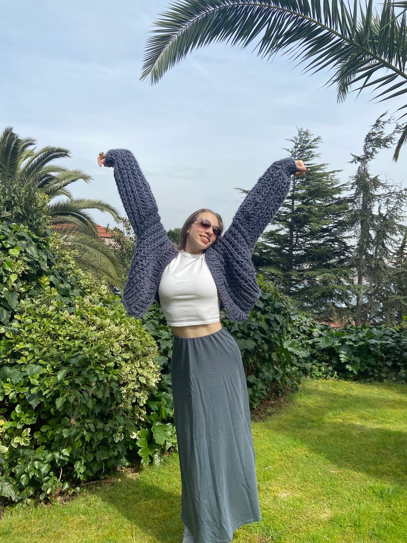 K&ouml;nnte beinhalten: Eine Frau tr&auml;gt einen grauen grob gestrickten Cardigan mit einem wei&szlig;en Crop-Top und einem langen grauen Rock. Sie steht in einem Garten mit Palmen im Hintergrund.