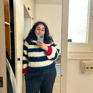 May include: A woman wearing a white and blue striped sweater with red and blue trim on the sleeves. She is wearing black pants and is taking a selfie.