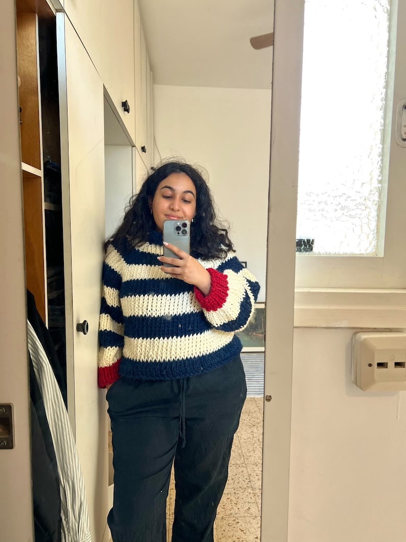 May include: A woman wearing a white and blue striped sweater with red and blue trim on the sleeves. She is wearing black pants and is taking a selfie.