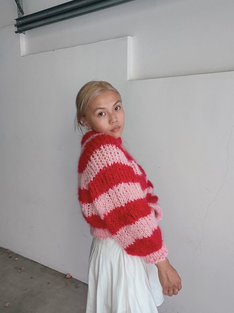 May include: A woman wearing a pink and red striped fuzzy sweater with a white skirt.