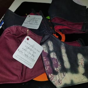 May include: A set of four face masks with a burgundy and black color scheme. The masks are made of fabric and have a printed design. The masks are accompanied by a white care tag with instructions for washing and drying the masks. The care tag reads "Machine wash gentle or hand wash line dry only".