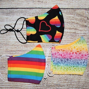 May include: Three colorful face masks with different patterns. The first mask has a black background with rainbow stripes and red hearts. The second mask has a rainbow striped pattern. The third mask has a yellow background with white floral designs.