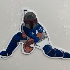 May include: A blue and white baseball player wearing a helmet with a red and white design. The player is in a catching position with a baseball in their glove. The player's uniform has the letters "LA" on the chest.