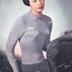 Op de afbeelding: Een vrouw draagt een lichtgrijze geribbelde trui met een klein zakje aan de linkerkant. De trui heeft een ronde hals en lange mouwen. De vrouw heeft kort, donker haar en draagt een donkere rok.