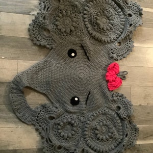 May include: A gray crocheted elephant rug with pink flower accents. The rug is made with a circular design and has a textured surface.