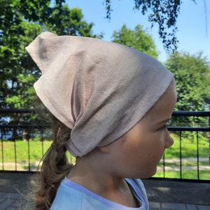 Peut inclure: Une jeune personne portant un bandana brun clair noué autour de la tête. Le bandana est fait d'un tissu doux et texturé. La personne porte un t-shirt tie-dye bleu clair et rose.