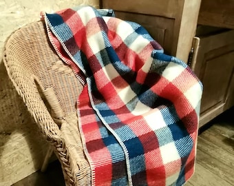 May include: A red, white, and blue plaid blanket draped over a wicker chair. The blanket has a woven texture and is folded over the back of the chair.