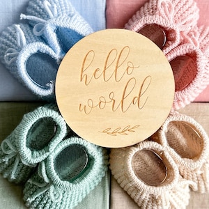 May include: A wooden round sign with the text "hello world" engraved on it. The sign is surrounded by four pairs of knitted baby booties in different colors: light blue, green, cream, and pink.