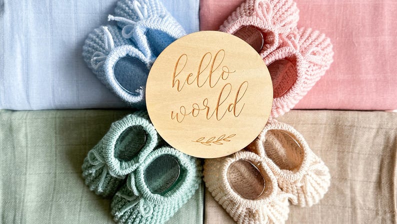 May include: A wooden round sign with the text "hello world" engraved on it. The sign is surrounded by four pairs of knitted baby booties in different colors: light blue, green, cream, and pink.