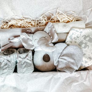 Puede incluir: Una caja de regalo para beb&eacute; de color gris y blanco llena de art&iacute;culos, incluyendo un juguete de peluche de elefante, un gorro de punto, botines y una manta.