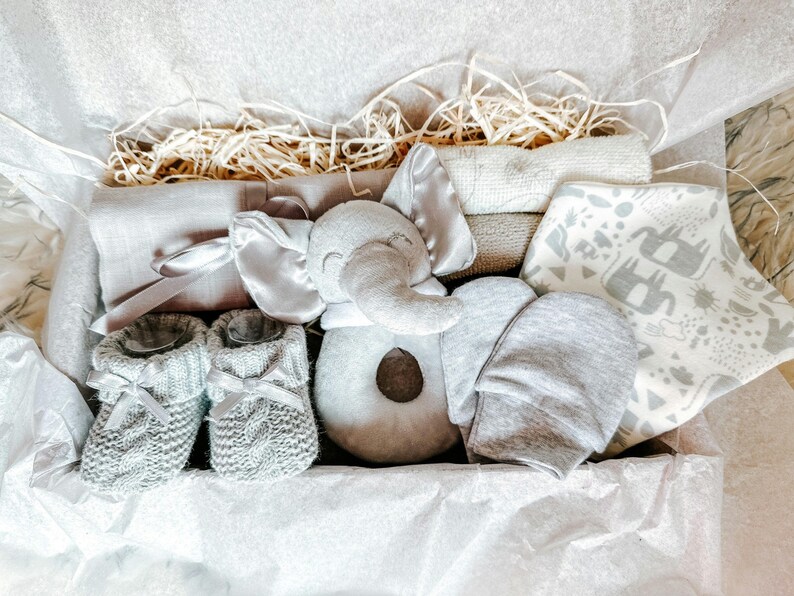Puede incluir: Una caja de regalo para beb&eacute; de color gris y blanco llena de art&iacute;culos, incluyendo un juguete de peluche de elefante, un gorro de punto, botines y una manta.