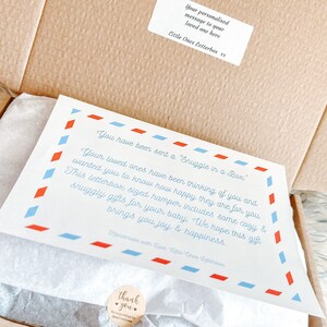 Puede incluir: Una tarjeta blanca con bordes azules y rojos y el texto "You have been sent a Snuggle in a Box." La tarjeta est&aacute; dentro de una caja de cart&oacute;n marr&oacute;n con papel de seda blanco. La tarjeta dice "Your loved ones have been thinking of you and wanted you to know how happy they are for you. This letterbox sized hamper includes some cozy & snuggly gifts for your baby. We hope this gift brings you joy & happiness."
