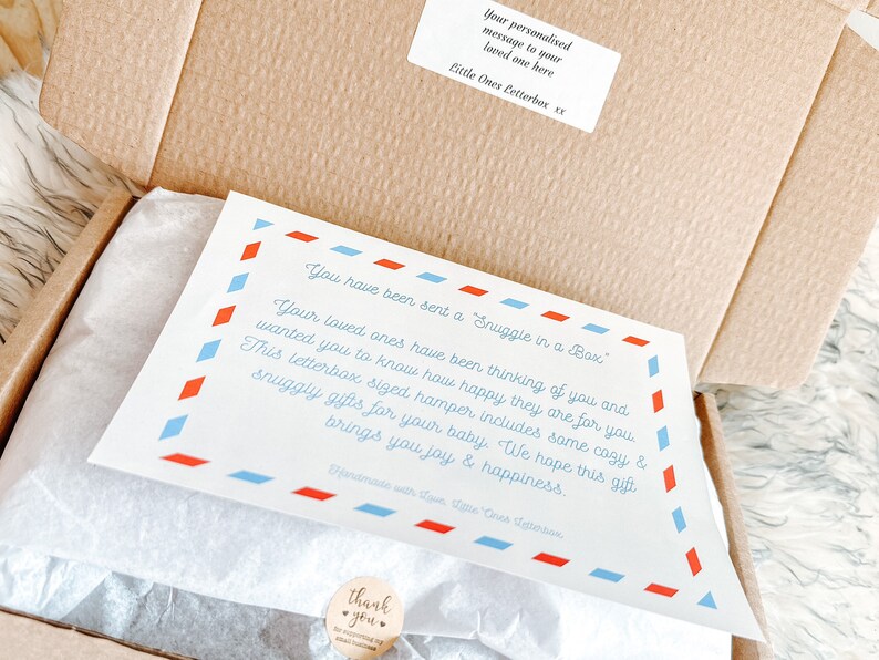 Puede incluir: Una tarjeta blanca con bordes azules y rojos y el texto "You have been sent a Snuggle in a Box." La tarjeta est&aacute; dentro de una caja de cart&oacute;n marr&oacute;n con papel de seda blanco. La tarjeta dice "Your loved ones have been thinking of you and wanted you to know how happy they are for you. This letterbox sized hamper includes some cozy & snuggly gifts for your baby. We hope this gift brings you joy & happiness."