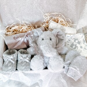 Puede incluir: Una caja de regalo para beb&eacute; en gris y blanco con un juguete de peluche de elefante, un par de botines de punto, una manopla de punto gris y una manta estampada en blanco y gris.