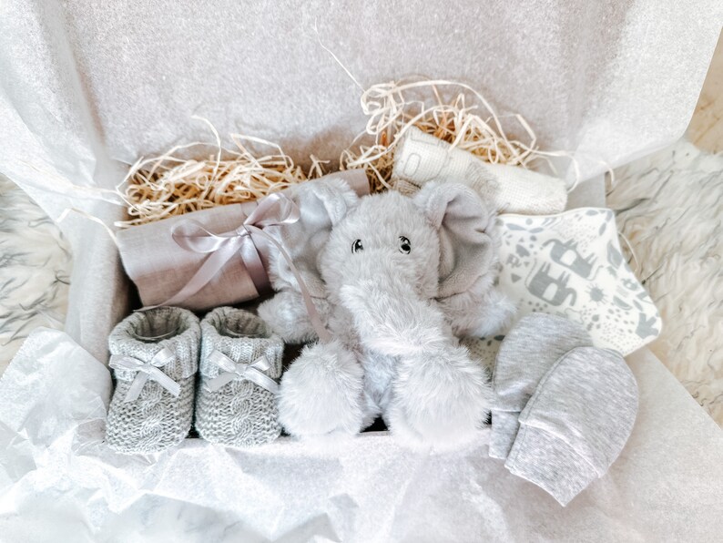 Puede incluir: Una caja de regalo para beb&eacute; en gris y blanco con un juguete de peluche de elefante, un par de botines de punto, una manopla de punto gris y una manta estampada en blanco y gris.