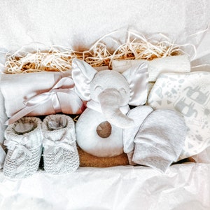Puede incluir: Un conjunto de regalo para beb&eacute; gris y blanco en una caja con papel de seda. El conjunto incluye un sonajero de elefante gris, un par de botines de punto grises, un babero estampado gris y blanco, un gorro gris y una manta gris y blanca.