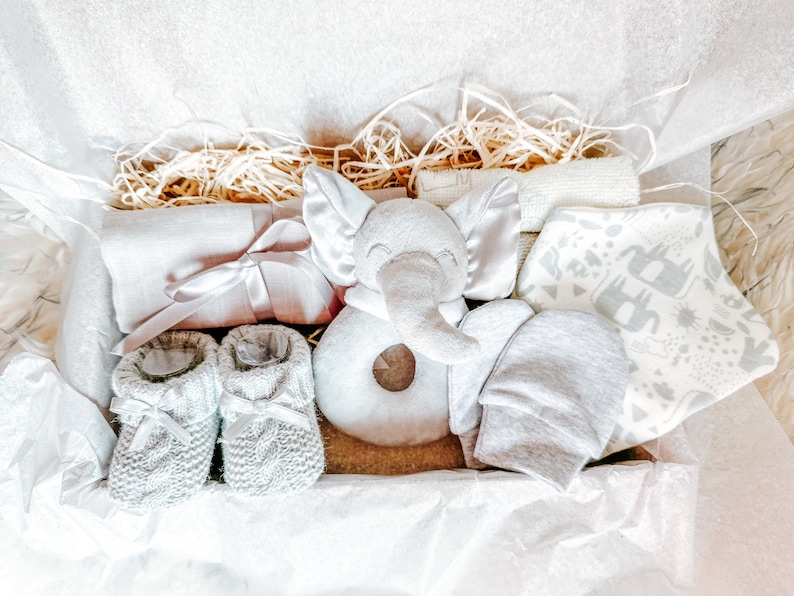 Puede incluir: Un conjunto de regalo para beb&eacute; gris y blanco en una caja con papel de seda. El conjunto incluye un sonajero de elefante gris, un par de botines de punto grises, un babero estampado gris y blanco, un gorro gris y una manta gris y blanca.