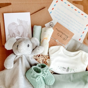 May include: A gift box filled with baby items, including a gray elephant plush toy, a white onesie with a green house design, green knitted booties, and a card that says "Congratulations Mummy to be".
