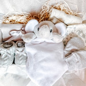 Puede incluir: Una cesta de regalo para beb&eacute; gris y blanca con un juguete de peluche de elefante, botines, una manta y un babero.