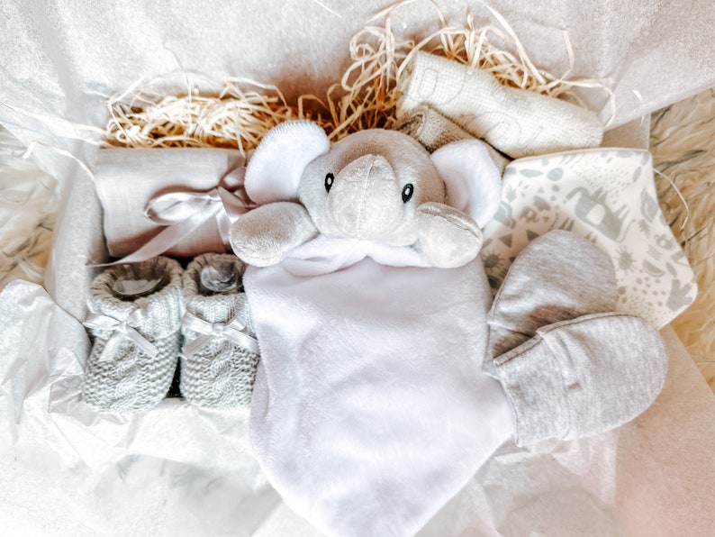 Puede incluir: Una cesta de regalo para beb&eacute; gris y blanca con un juguete de peluche de elefante, botines, una manta y un babero.