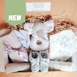 Puede incluir: Una caja de regalo llena de art&iacute;culos para beb&eacute;s, que incluyen un sonajero de elefante gris, un babero estampado en gris y blanco, una botita de beb&eacute; de punto gris y una muselina gris. La caja est&aacute; forrada con papel marr&oacute;n y paja.