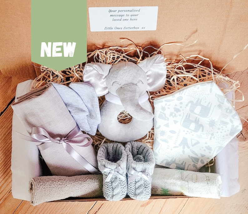 Puede incluir: Una caja de regalo llena de art&iacute;culos para beb&eacute;s, que incluyen un sonajero de elefante gris, un babero estampado en gris y blanco, una botita de beb&eacute; de punto gris y una muselina gris. La caja est&aacute; forrada con papel marr&oacute;n y paja.