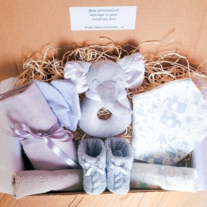 Puede incluir: Una caja de regalo llena de art&iacute;culos para beb&eacute;s, que incluyen un sonajero de elefante gris, un babero estampado en azul y blanco, un par de botines de punto azul y una muselina gris y blanca. La caja est&aacute; forrada con papel triturado y tiene una etiqueta que dice "Tu mensaje personalizado a tu ser querido aqu&iacute; Little Ones Letterbox xx".