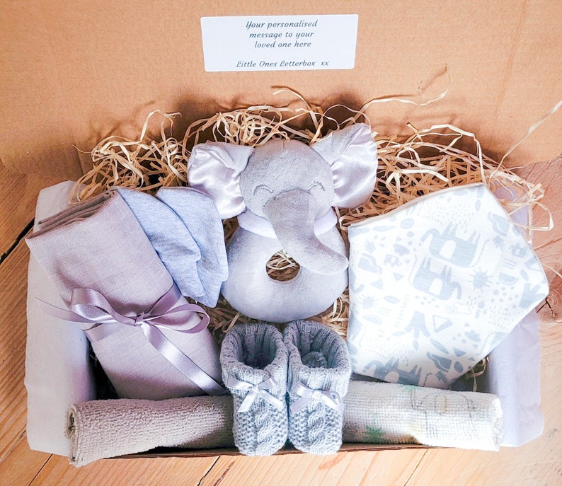 Puede incluir: Una caja de regalo llena de art&iacute;culos para beb&eacute;s, que incluyen un sonajero de elefante gris, un babero estampado en azul y blanco, un par de botines de punto azul y una muselina gris y blanca. La caja est&aacute; forrada con papel triturado y tiene una etiqueta que dice "Tu mensaje personalizado a tu ser querido aqu&iacute; Little Ones Letterbox xx".